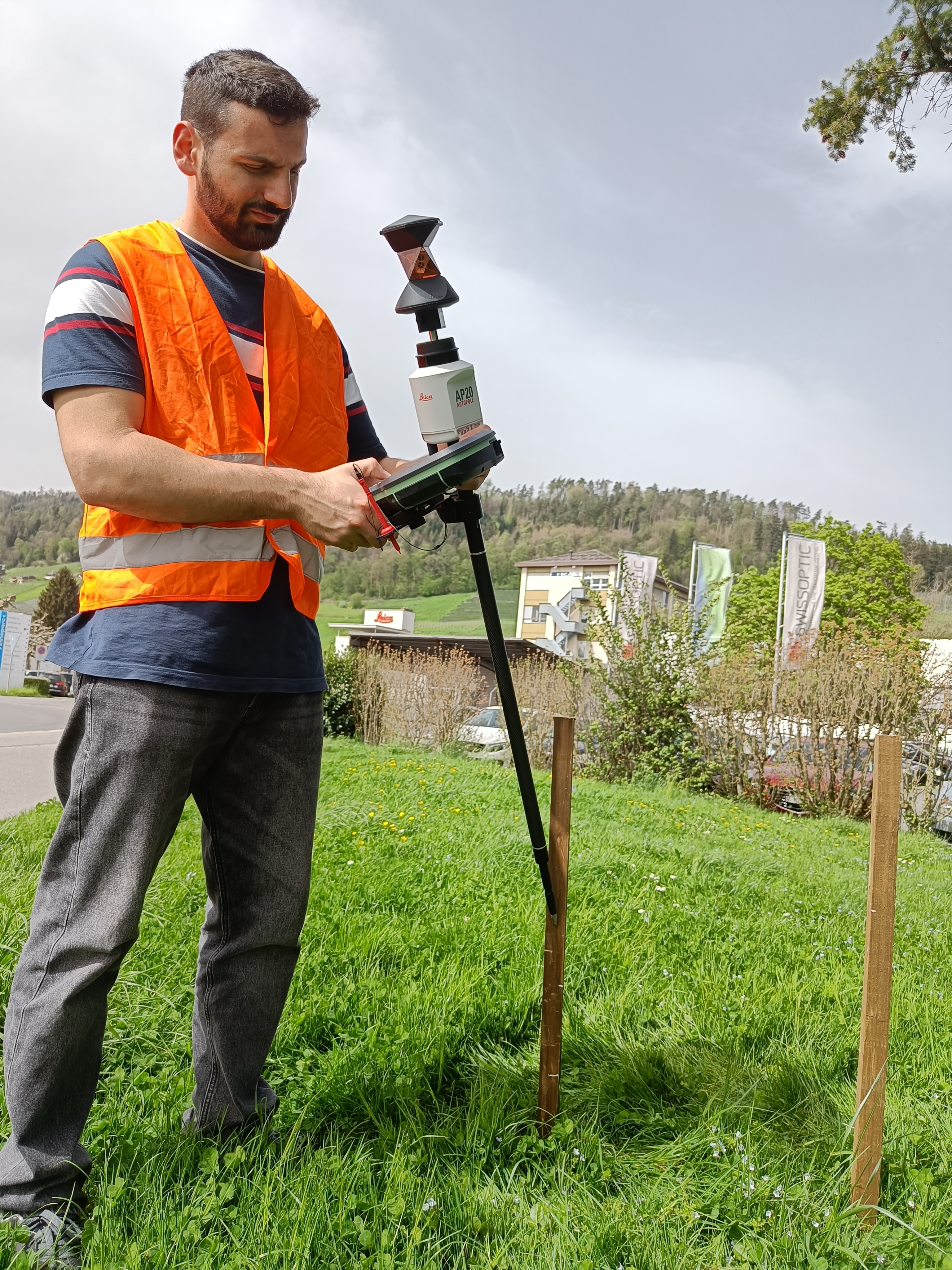 A surveyor in an orange safety vest operates a state-of-the-art Leica Geosystems AP20 AutoPole in a grassy area with hills and buildings in the background.